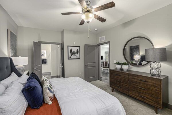 Bedroom with carpet, plush bed with headboard, dresser with lamp and decor, and framed mirror.