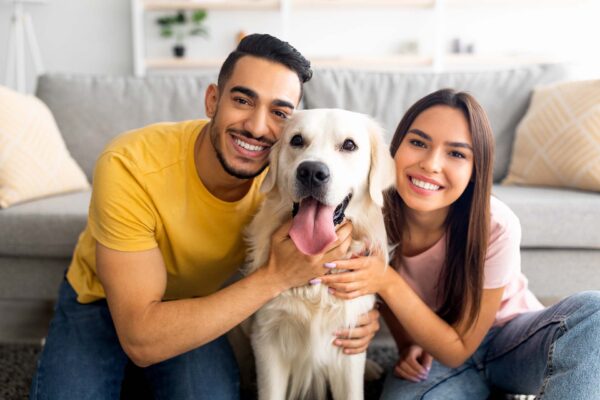 Portrait of smiling diverse couple hugging their pet dog, smiling at camera, indoors. Arab guy and his Caucasian girlfriend embracing their golden retriever at home
