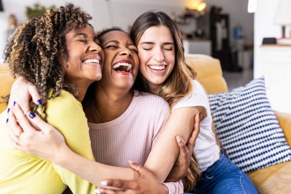 Three happy multiethnic female best friends laughing together - Diverse young women hugging each other and having fun at home - Unity concept