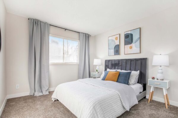 Kenilworth apartment bedroom with carpet, plush bed with headboard, bedside tables with lamps, and large window.