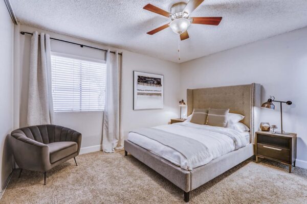 Latitude apartment bedroom with carpet, plush bed with headboard, bedside tables with lamps, and large window.