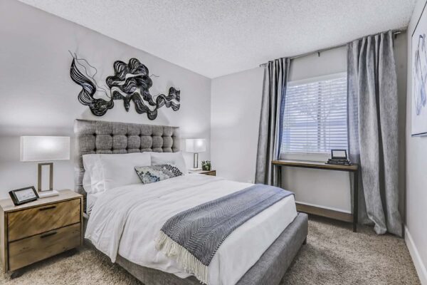 Latitude apartment bedroom with carpet, plush bed with headboard, bedside tables with lamps, and large window.