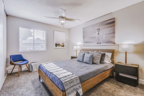Latitude apartment bedroom with plush carpet, wood bed with headboard, modern chair, and ceiling fan.