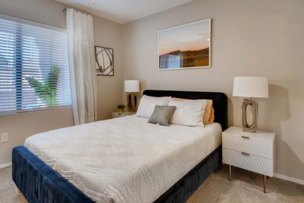 Mandarina apartment bedroom with carpet, plush bed with headboard, bedside tables with lamps, and large window.