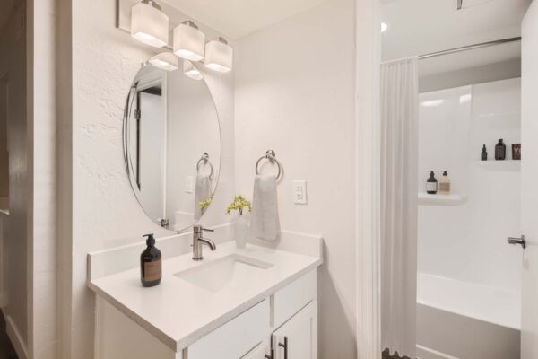 Omnia on Thomas apartment bathroom with white cabinets and counters, round mirror, and tiled shower tub.