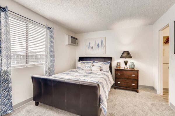 Overlook at Sunset Point apartment bedroom with carpet, plush bed with headboard, bedside drawer with lamp, and large window.