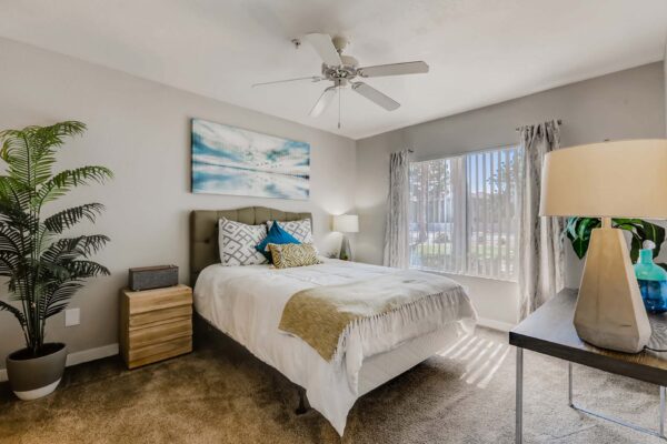 Pavilions at Arrowhead apartment bedroom with carpet, plush bed with headboard, large potted plant, and large window.