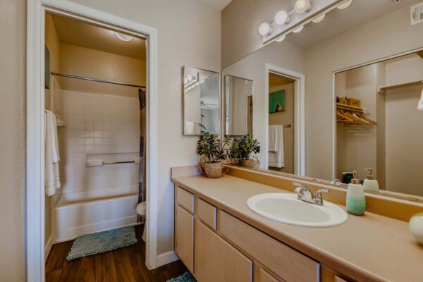Pavilions at Arrowhead apartment bathroom with wood style floor, tan cabinets and counter, and large mirror.