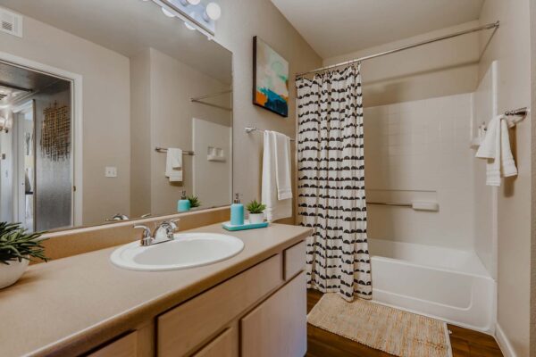 Pavilions at Arrowhead apartment bathroom with wood style floor, tan cabinets and counters, and tiled shower tub.