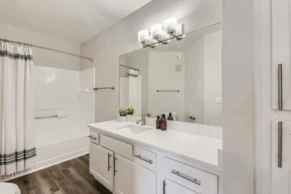 Pavilions at Arrowhead apartment bathroom with wood style floor, white cabinets and counters, and tiled shower tub.
