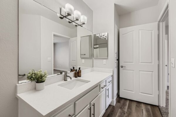 Pavilions at Arrowhead apartment bathroom with wood style floor, white cabinets and counters, large mirror, and designer lights.