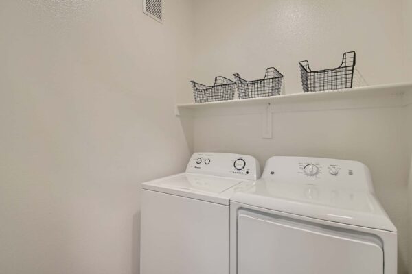 Springs at Alta Mesa apartment laundry room with washer, dryer, and shelf with baskets.