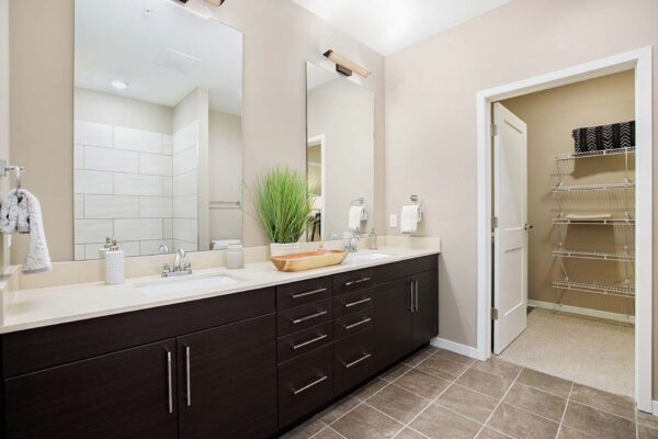 The Locale apartment bathroom with tile floor, dark cabinets, light counters, dual mirrors, and large closet.
