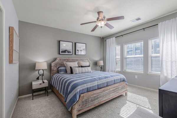 Village West apartment bedroom with carpet, plush bed with headboard, bedside tables with lamps, and large windows.