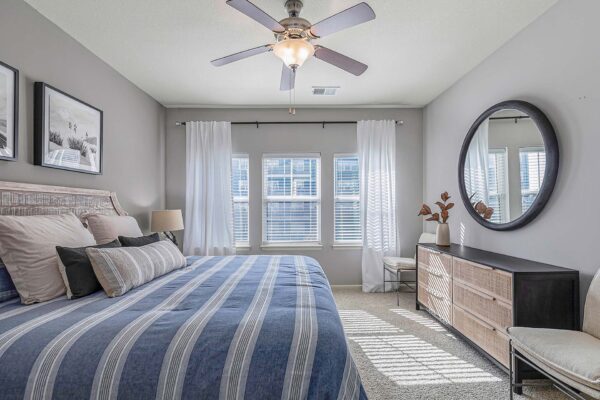 Village West apartment bedroom with carpet, plush bed with headboard, dresser, and large round mirror.
