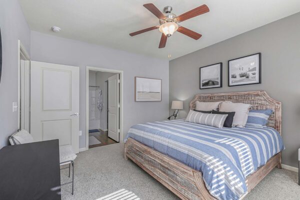 Village West apartment bedroom with carpet, plush bed with headboard, framed artwork, and bathroom door.