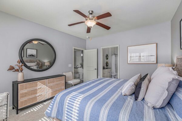 Village West apartment bedroom with carpet, plush bed, dresser, and large round mirror.