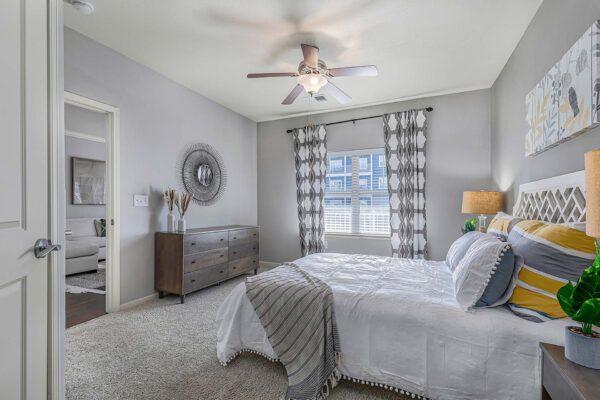 Village West apartment bedroom with carpet, plush bed with headboard, bedside tables with lamps, and ceiling fan.