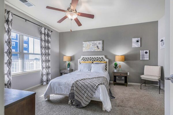 Village West apartment bedroom with carpet, plush bed with headboard, bedside tables with lamps, and ceiling fan.