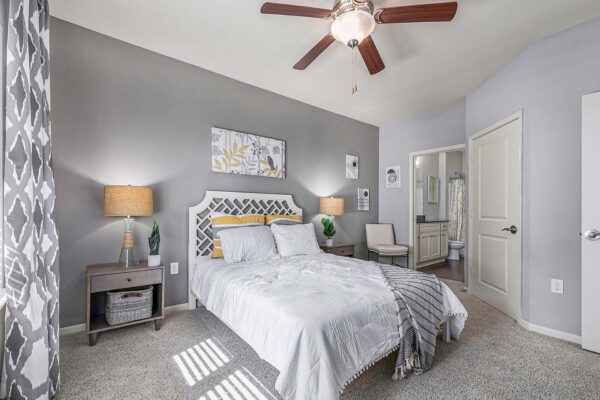 Village West apartment bedroom with carpet, plush bed with headboard, bedside tables with lamps, and ceiling fan.