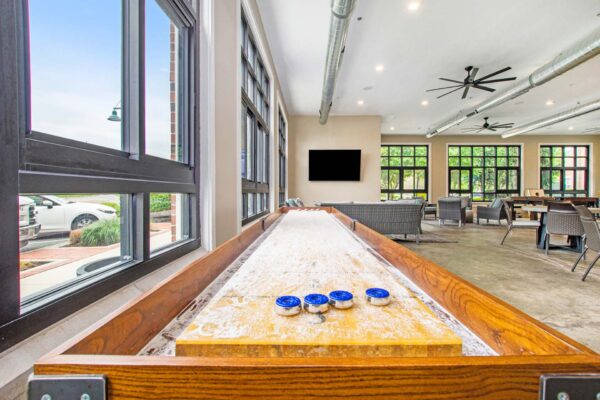Village at Mission Farms apartments community lounge with shuffleboard table and large windows.