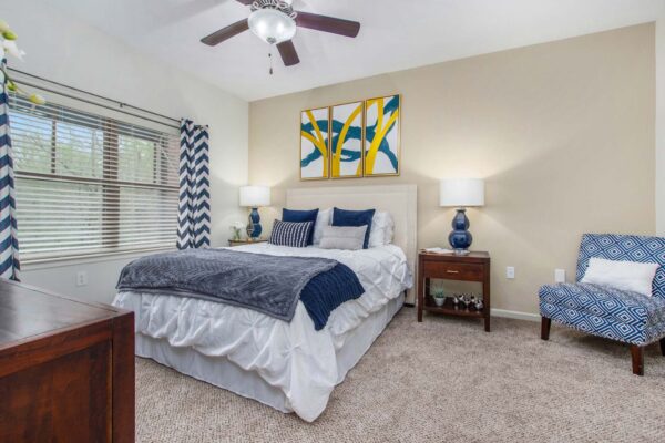 Village at Mission Farms apartment bedroom with carpet, plush bed with headboard, bedside tables with lamps, and ceiling fan.