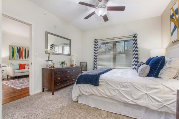 Village at Mission Farms apartment bedroom with carpet, plush bed with headboard, wood dresser, framed mirror, and ceiling fan.