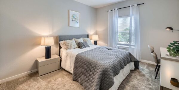 The Avery apartment bedroom with carpet, plush bed with headboard, desk, and large window.