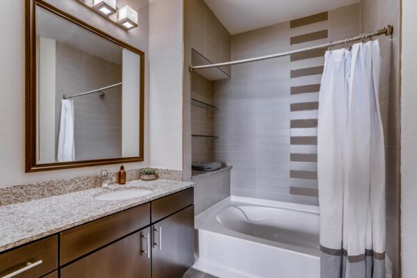 Centre Court apartment bathroom with wood cabinets, stone counter, framed mirror, and tiled shower tub.