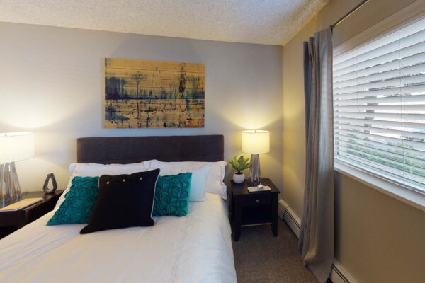 Flats at Pinecliff apartment bedroom with carpet, plush bed with headboard, and large window with curtain.