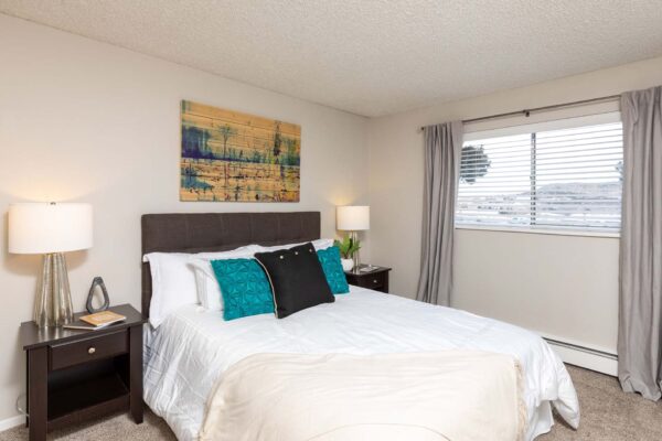 Flats at Pinecliff apartment bedroom with carpet, plush bed with headboard, large windows, and artwork.