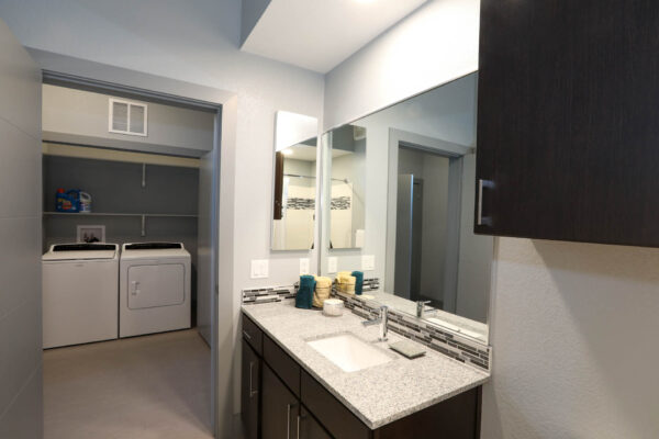 Forum Fitzsimons apartment bathroom with dark cabinets, light counters, large mirror, and laundry room door.