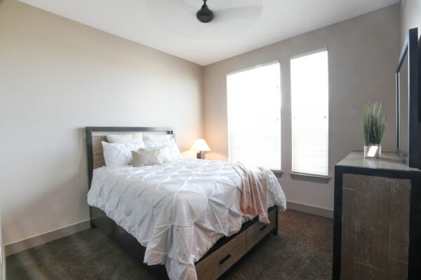 Forum Fitzsimons apartment bedroom with carpet, plush bed with headboard, dresser, and large window.