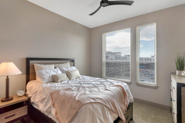 Forum Fitzsimons apartment bedroom with carpet, plush bed with headboard, bedside table with lamp, and large window.