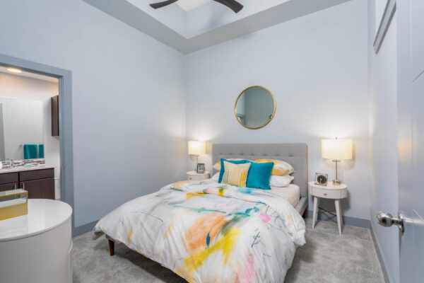 Forum Fitzsimons apartment bedroom with carpet, plush bed with headboard, bedside tables with lamps, and large mirror.