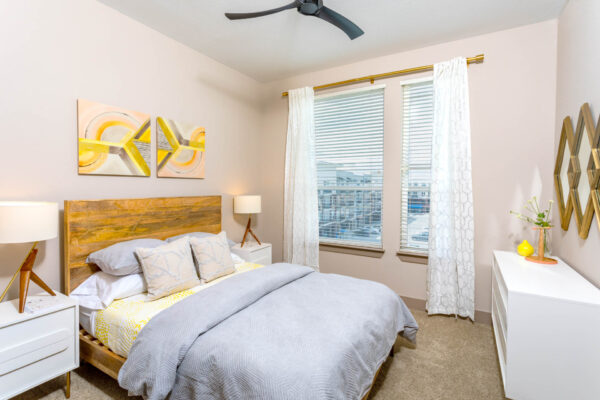 Forum Fitzsimons apartment bedroom with carpet, plush bed with headboard, bedside tables with lamps, and large windows.