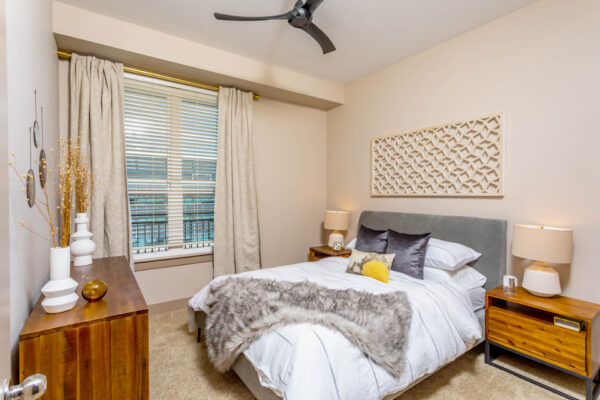 Forum Fitzsimons apartment bedroom with carpet, plush bed with headboard, bedside tables with lamps, and large window.