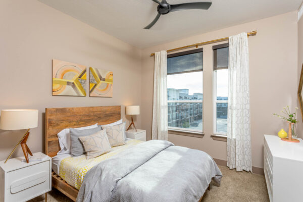 Forum Fitzsimons apartment bedroom with carpet, plush bed with headboard, bedside drawers with lamps, and large windows.