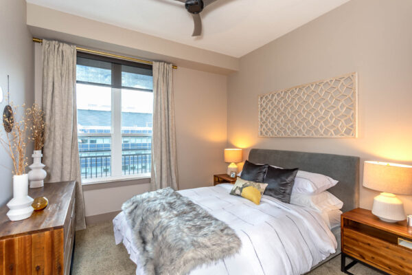 Forum Fitzsimons apartment bedroom with carpet, plush bed with headboard, bedside tables with lamps, and large window.