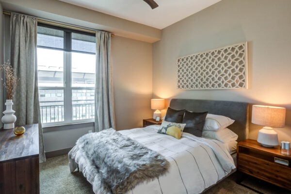 Forum Fitzsimons apartment bedroom with carpet, plush bed with headboard, bedside tables with lamps, and large windows.