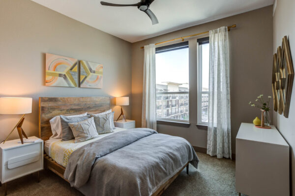 Forum Fitzsimons apartment bedroom with carpet, plush bed with headboard, bedside tables with lamps, and large windows.