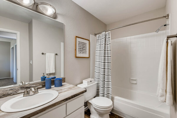 Sunset Peak apartment bathroom with white cabinets, stone counters, large mirror, and tiled shower tub.