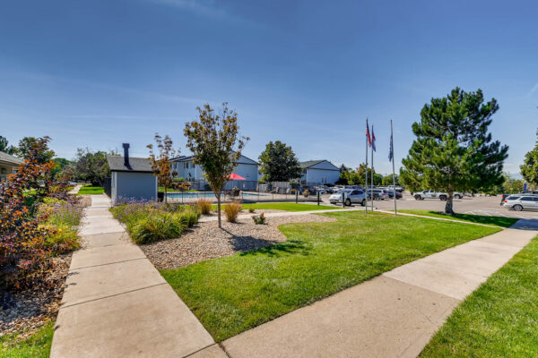 Sunset Peak Apartments outdoor scene featuring a landscaped pathway, grassy areas, a swimming pool in the background, and nearby flagpoles.