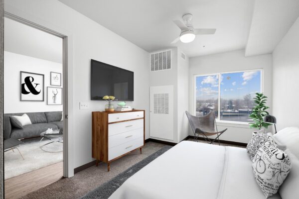 The Overland apartment bedroom with carpet, plush bed, dresser, TV, and large window.