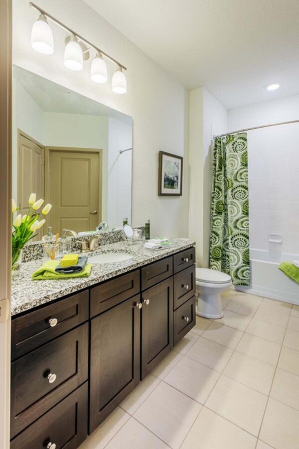 The Point at Bella Grove apartments bathroom with tiled floors, dark wood cabinets, quartz counters, tiled shower tub, lighting fixtures and large mirror.
