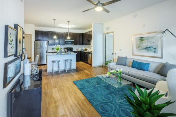 The Point at Bella Grove apartments living room with wood style floor, plush seating, ceiling fan and show casing modern kitchen.