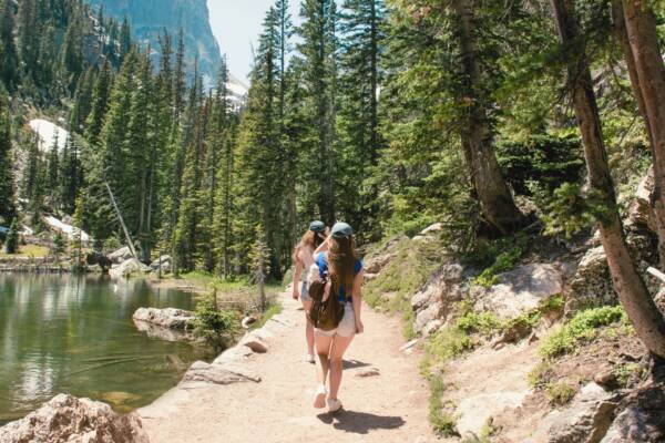 People hiking alongside mountain lake under tall pine trees in Colorado.