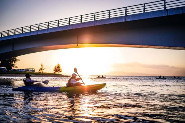 People kayaking under a bridge at sunset.