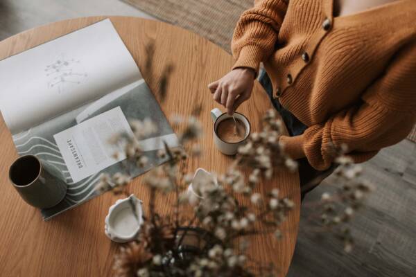 Person mixing a latte and reading from a large magazine at a wooden table.